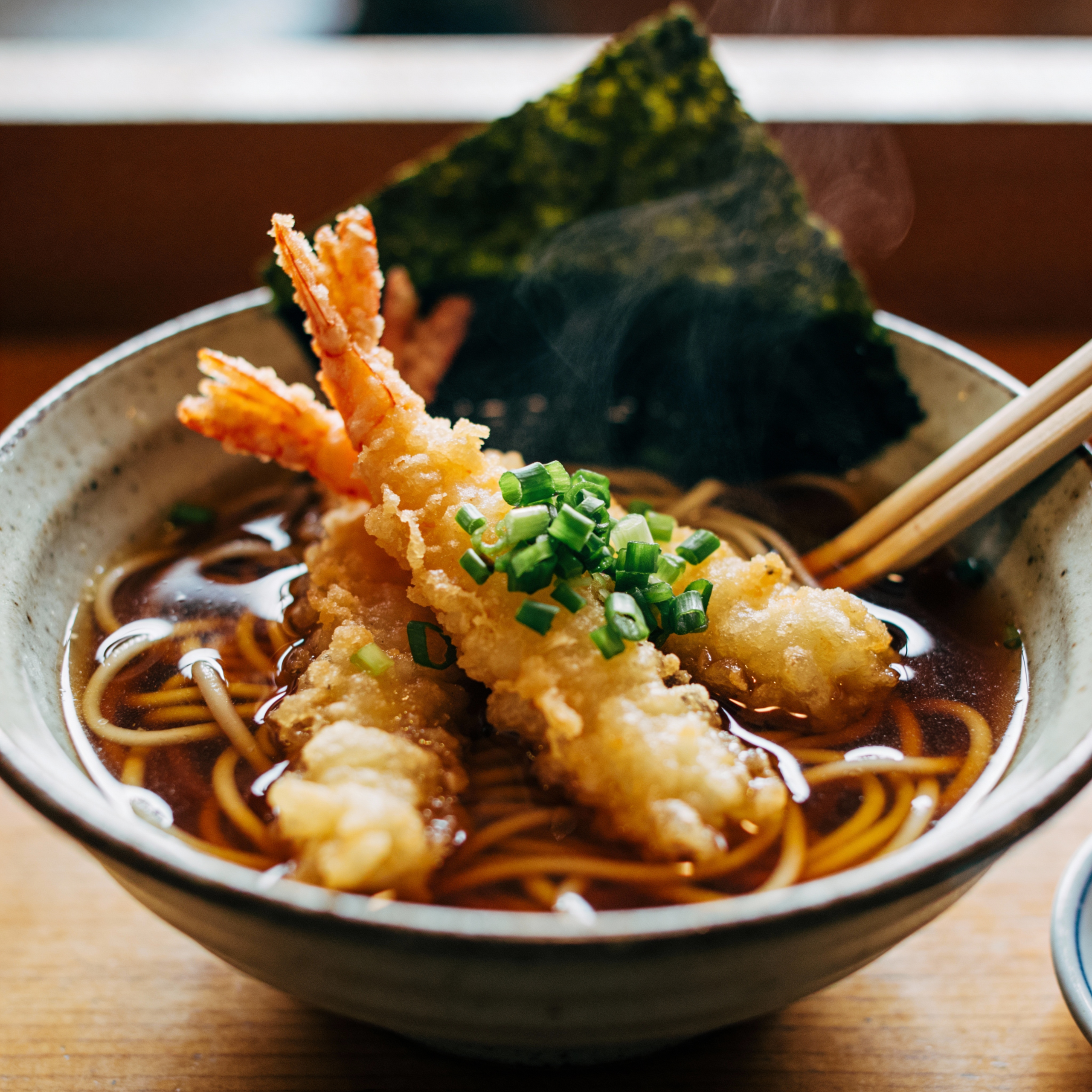 Close-up food photography of "Toshikoshi Soba" (Japanese New Year buckwheat noodles) in a ceramic bowl. Topped with tempura shrimp and chopped green onions. Steam rising from the hot broth. Warm, cozy lighting, eaten with chopsticks.