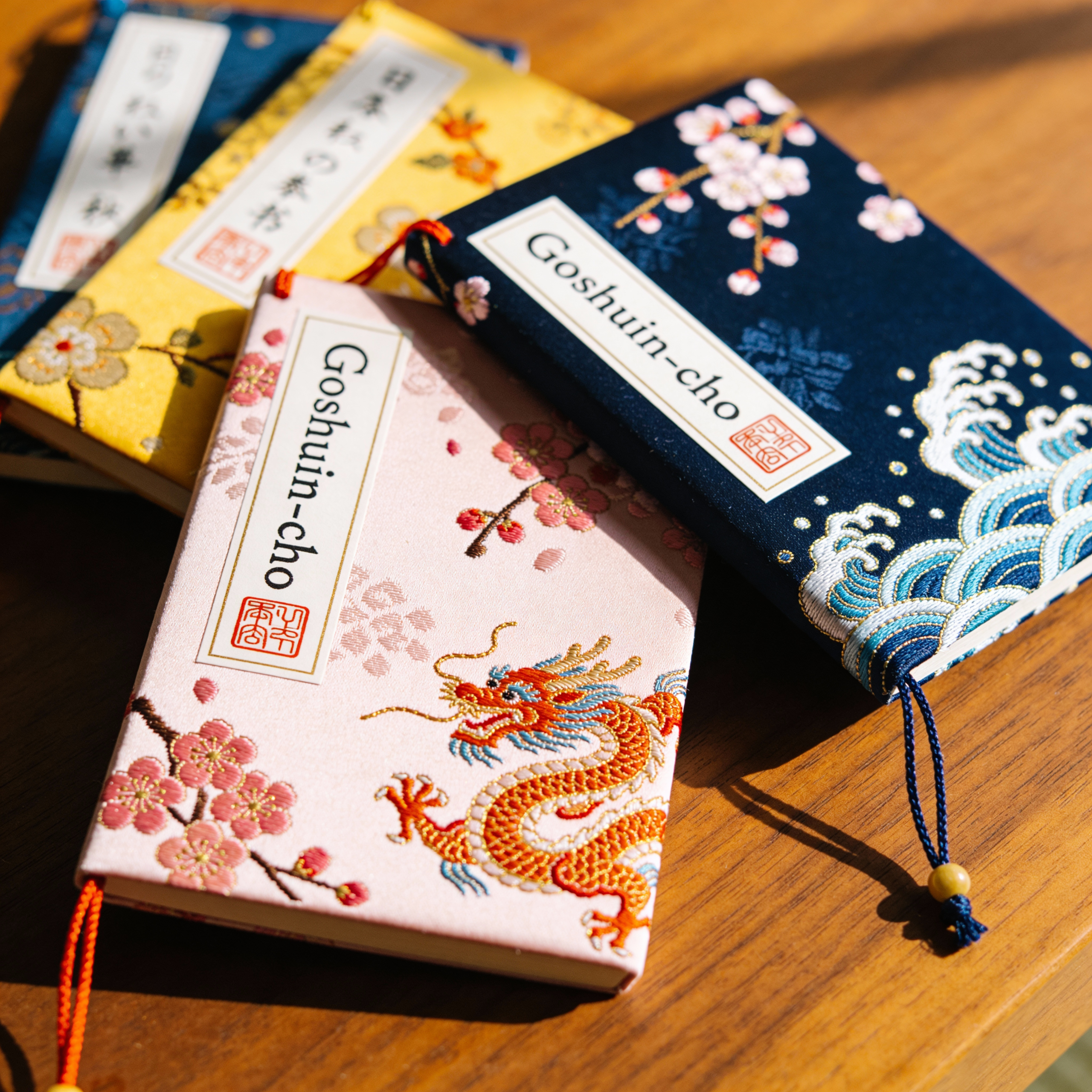 Product photography of several colorful "Goshuin-cho" (Japanese stamp books) lying on a wooden table. The covers feature beautiful traditional Japanese embroidery patterns like cherry blossoms, dragons, and waves. High quality, elegant stationery style.