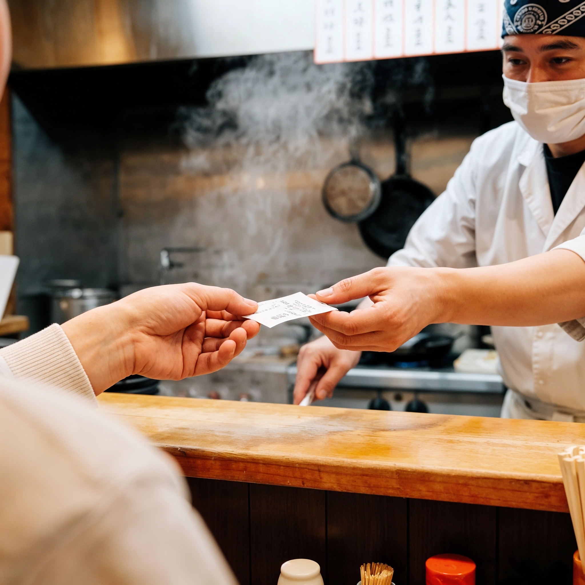 A candid shot from the customer's perspective across a wooden ramen counter. A hand is extending to give a small paper ticket to the hand of a busy Ramen chef (wearing a bandana or uniform) in the kitchen showing only his hands and part of his body. Steam rising in the background.