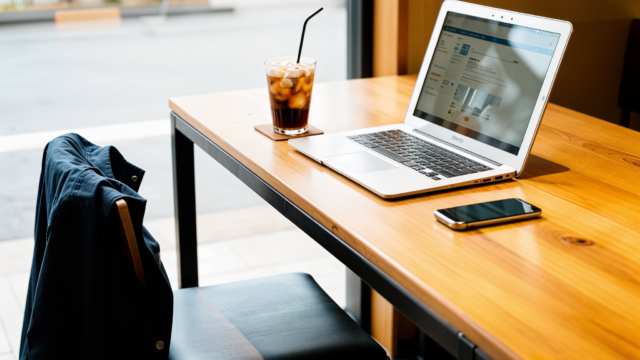 Photography of a table in a stylish Tokyo cafe. A laptop, a smartphone, and an iced coffee are left unattended on the wooden table. A jacket hangs on the chair. No one is sitting there. Background shows a blurred, peaceful cafe interior. Concept of safety and trust.